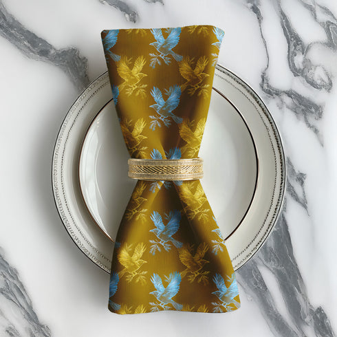 Set of four golden cloth napkins featuring alternating powder blue and metallic gold ravens in flight, sized 18x18 inches.